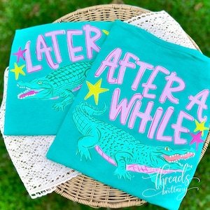 Peut inclure: Deux t-shirts turquoise avec des graphiques d'alligators et du texte. L'un dit "LATER" en rose, l'autre "AFTER A WHILE" en rose. Les deux chemises ont des étoiles jaunes et des accents roses.