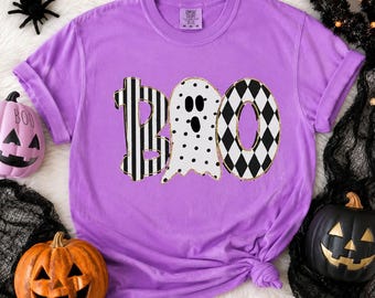 May include: A lavender short-sleeve t-shirt featuring the word BOO printed in large letters. The B has black and white vertical stripes, the O is a white ghost with black polka dots and facial features, and the second O has a black diamond pattern.