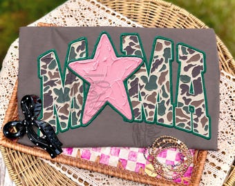 May include: A dark gray t-shirt featuring the word MAMA in large block letters with a camouflage pattern. The letter A is replaced by a textured light pink star. The letters are outlined in dark green. A black hair clip and beaded bracelets are nearby.