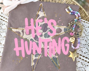 May include: A brown t-shirt featuring a large star with a camouflage pattern and the pink text HE'S HUNTING. The shirt is accented with a light pink bow at the top and a collection of colorful beaded bracelets with a gold star charm on the side.