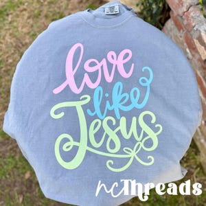 May include: A gray t-shirt with the words "love like Jesus" in pink, blue, and green script. The text is centered on the front of the shirt. The brand name "ncThreads" is at the bottom.