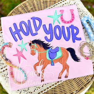 May include: Pink t-shirt with the words "HOLD YOUR" in blue, a cartoon horse, stars, and a horseshoe. The horse is brown with a blue saddle. Accessories like a necklace and hair clips are in the background.