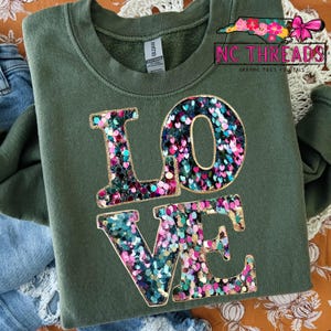 Könnte beinhalten: Olivgrünes Sweatshirt mit dem Wort "LOVE" in großen, glitzernden Buchstaben. Die Buchstaben sind mit einer Mischung aus rosa, türkis und goldenem Glitzer gefüllt. Das Sweatshirt hat einen Rundhalsausschnitt und lange Ärmel. Der Text "NC THREADS GRAPHIC TEES + DIGITALS" befindet sich oben rechts.