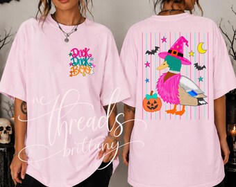 May include: A light pink t-shirt featuring a graphic on the front left chest that reads Duck Duck BOO in colorful text with small stars and a ghost. The back displays a mallard duck wearing a pink witch hat, a pumpkin, and stars.