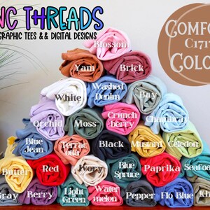 May include: A display of rolled-up t-shirts in various colors, including white, blue, red, and yellow. The image features the text "NC THREADS GRAPHIC TEES & DIGITAL DESIGNS" and "COMFORT C1717 COLORS".