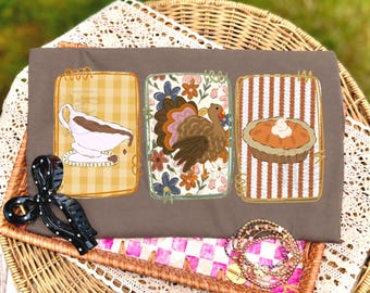May include: A brown t-shirt featuring three rectangular graphic panels. The left panel shows a gravy boat on a yellow gingham background. The center panel displays a turkey over a floral pattern. The right panel depicts a pumpkin pie on stripes.