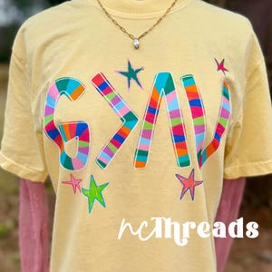 May include: A light yellow t-shirt with the word "G>A>V" in colorful, striped letters and star graphics. The text "ncThreads" is printed in white script at the bottom. A gold necklace with a pendant is visible.