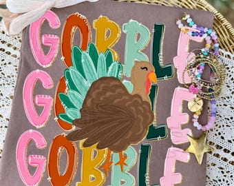 May include: A brown t-shirt featuring a graphic of a turkey and the text GOBBLE GOBBLE GOBBLE in pink, orange, yellow, and green. A light-colored ribbon is tied at the top left, and a stack of colorful beaded bracelets with a gold star charm sits on the right.