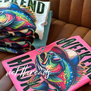 May include: A stack of t-shirts featuring a colorful graphic of a fish wearing sunglasses and a hat. The shirts are in white, teal, and pink, with the word "WEEKEND" printed on them in black and green.
