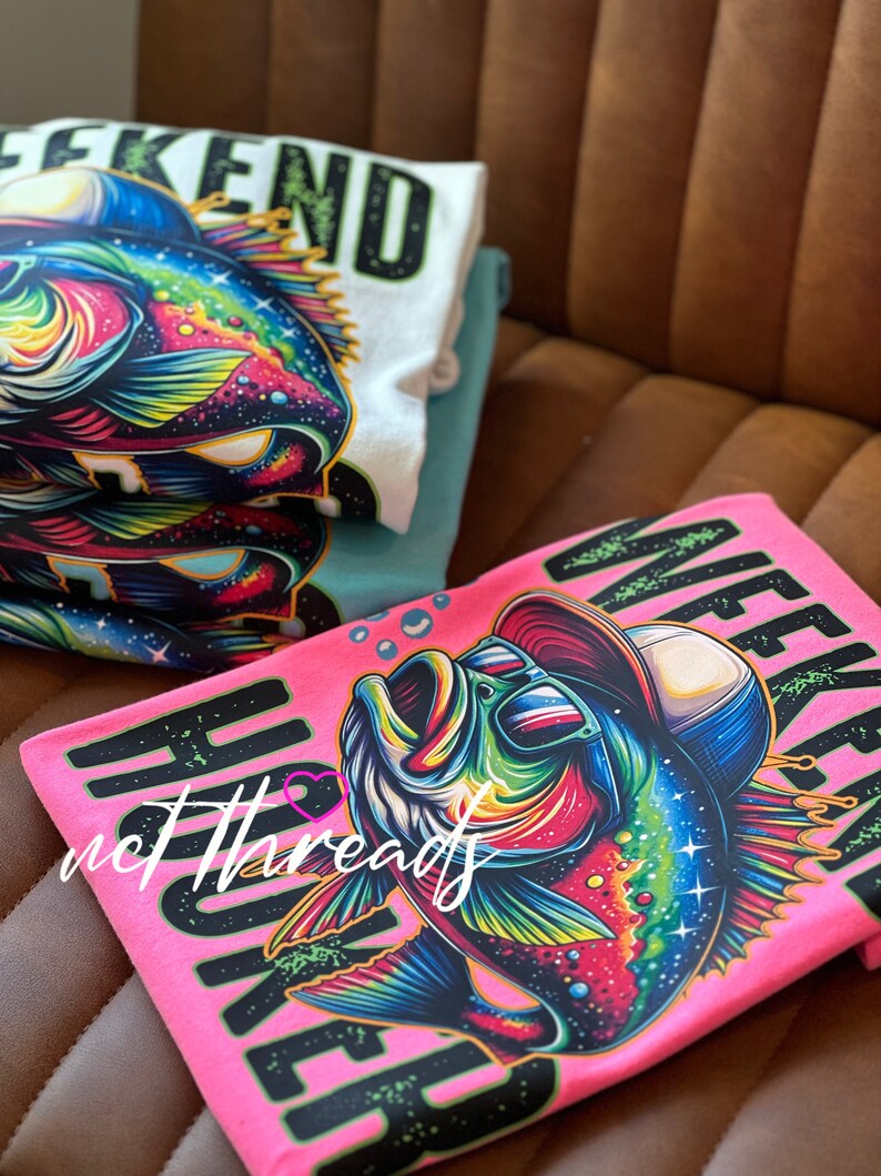 May include: A stack of t-shirts featuring a colorful graphic of a fish wearing sunglasses and a hat. The shirts are in white, teal, and pink, with the word "WEEKEND" printed on them in black and green.