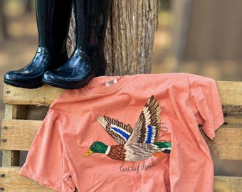 May include: A coral-colored short-sleeve t-shirt featuring an embroidered mallard duck in flight with brown, green, and blue details, accompanied by the cursive text "lucky duck" below the bird, paired with a pair of black Hunter rain boots.