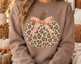 May include: A brown long-sleeve sweatshirt featuring a large graphic of a pumpkin with a leopard print pattern. The pumpkin is decorated with a brown and white gingham check bow tied at the top. The sweatshirt has a crew neckline and ribbed cuffs.