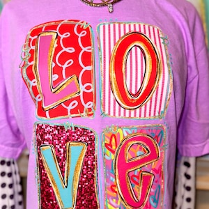 Puede incluir: Una camiseta lila con la palabra "LOVE" en un diseño colorido de letras en bloque. Las letras están en cuadrados individuales con diferentes patrones, incluyendo remolinos rojos, rayas rosas y purpurina. La camiseta tiene mangas cortas.