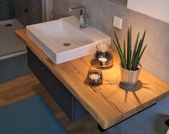 May include: A white porcelain bathroom sink with a chrome faucet sits on a wooden countertop with a gray cabinet underneath. Two glass candle holders and a potted plant are on the countertop.
