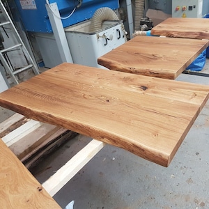 May include: Three unfinished wooden tabletops with live edges. The tabletops are made of light brown wood with a natural grain pattern. The tabletops are stacked on top of each other, with the largest tabletop on the bottom and the smallest tabletop on top.