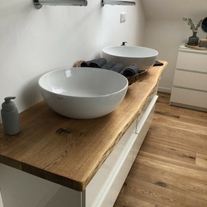 May include: White bathroom vanity with two white porcelain sinks, a wooden countertop, and white drawers. The countertop is a live edge slab of wood.