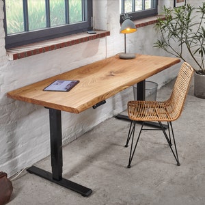 Peut inclure: Un bureau en bois avec un plateau à bord vif et des pieds réglables noirs. Une chaise en osier se trouve à côté du bureau. Une lampe avec un abat-jour gris est posée sur le bureau.