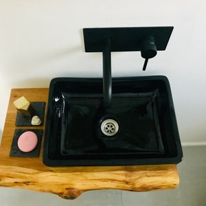 May include: A black ceramic bathroom sink with a black faucet mounted on a wooden countertop. The countertop has three small black square trays with soap and a pink bar of soap.