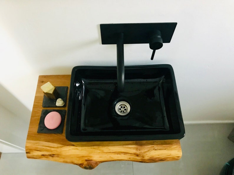 May include: A black ceramic bathroom sink with a black faucet mounted on a wooden countertop. The countertop has three small black square trays with soap and a pink bar of soap.