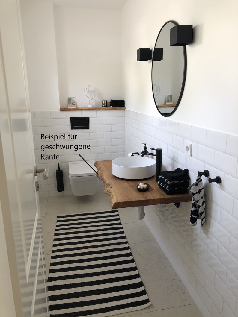 May include: A white bathroom with white subway tile walls, a black and white striped rug, a white toilet, a white sink with a wooden countertop, and a round black mirror with black sconces.