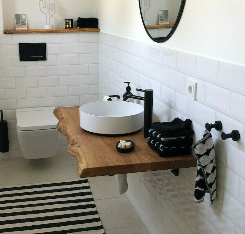 May include: A white porcelain sink with a black faucet mounted on a wooden countertop. The countertop is a natural edge slab with a live edge. Black towels are folded on the countertop and a black and white patterned towel hangs on a hook.