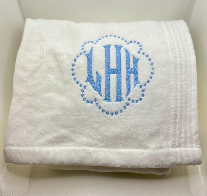 May include: A folded white towel with a light blue monogram. The monogram features the letters LHH inside a scalloped border. The towel has a plush texture and a ribbed border.