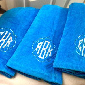 May include: Three folded turquoise towels with white monogrammed initials. Each towel features a different set of initials within a decorative, scalloped border. The towels are made of a soft, plush material.