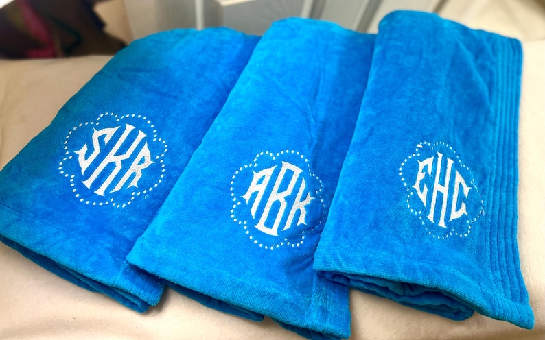 May include: Three folded turquoise towels with white monogrammed initials. Each towel features a different set of initials within a decorative, scalloped border. The towels are made of a soft, plush material.