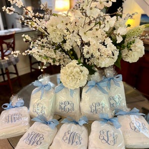 May include: White monogrammed gifts wrapped in clear cellophane and tied with blue gingham ribbons. A large bouquet of white flowers sits in the background. The gifts are arranged on a glass table.