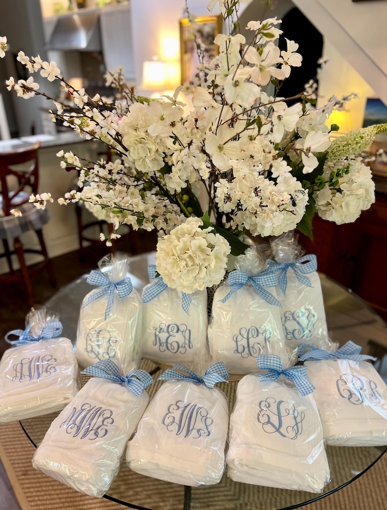 May include: White monogrammed gifts wrapped in clear cellophane and tied with blue gingham ribbons. A large bouquet of white flowers sits in the background. The gifts are arranged on a glass table.