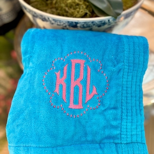 May include: A folded turquoise towel with a pink monogram. The monogram is in a decorative, floral-like frame. The towel has a ribbed texture on the right side.