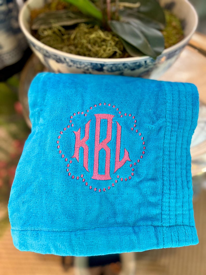 May include: A folded turquoise towel with a pink monogram. The monogram is in a decorative, floral-like frame. The towel has a ribbed texture on the right side.