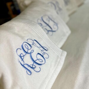 May include: White plush blankets with embroidered blue initials. The blankets are folded and stacked, showcasing the detailed monogram design. The soft texture and elegant embroidery create a luxurious feel.