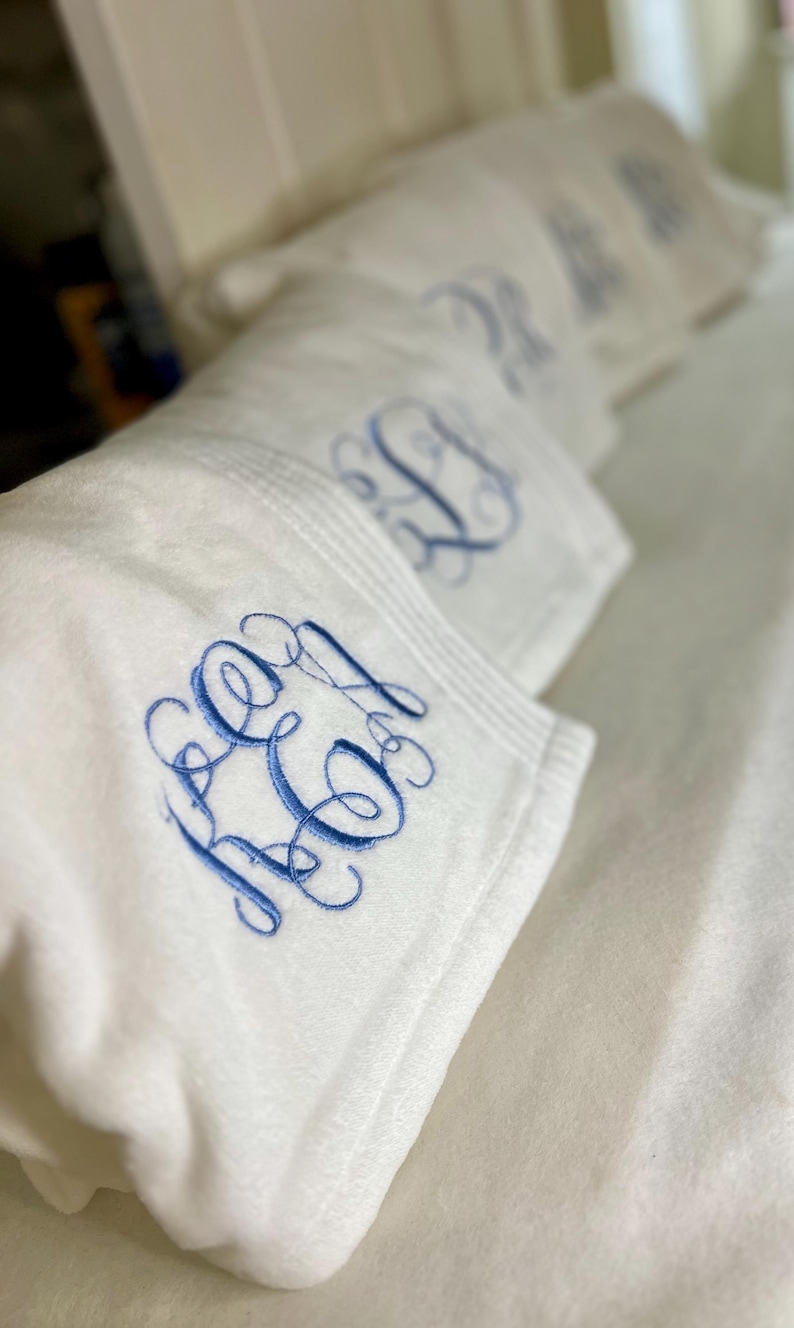 May include: White plush blankets with embroidered blue initials. The blankets are folded and stacked, showcasing the detailed monogram design. The soft texture and elegant embroidery create a luxurious feel.