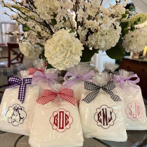 May include: White gift bags with monogrammed designs, tied with gingham and solid color bows. The bags are displayed in front of a large arrangement of white flowers, creating a decorative and elegant presentation.