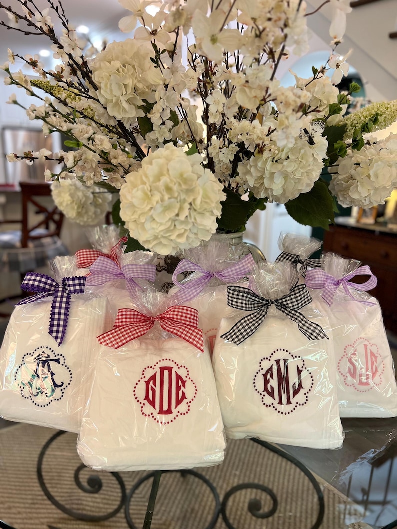 May include: White gift bags with monogrammed designs, tied with gingham and solid color bows. The bags are displayed in front of a large arrangement of white flowers, creating a decorative and elegant presentation.