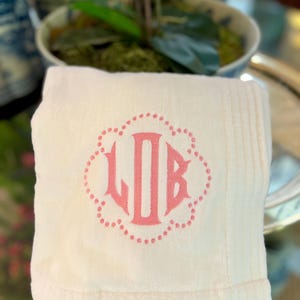 May include: A folded, cream-colored towel with a pink monogram of the letters LJB inside a dotted floral design. The towel has a soft, plush texture and a ribbed border. The background is blurred, suggesting a home decor setting.