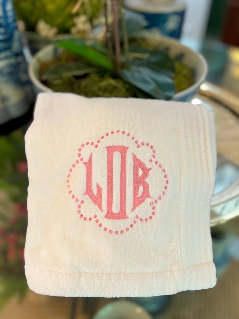 May include: A folded, cream-colored towel with a pink monogram of the letters LJB inside a dotted floral design. The towel has a soft, plush texture and a ribbed border. The background is blurred, suggesting a home decor setting.