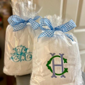 May include: Two white towels, each wrapped in clear cellophane and tied with a blue and white gingham ribbon. One towel has a teal monogram, the other has a green and purple monogram.
