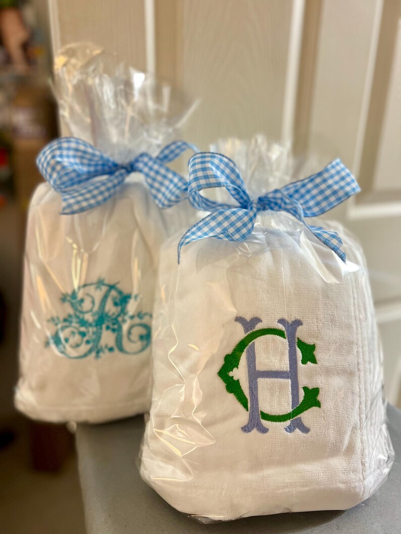 May include: Two white towels, each wrapped in clear cellophane and tied with a blue and white gingham ribbon. One towel has a teal monogram, the other has a green and purple monogram.