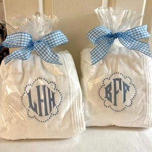 May include: Two white monogrammed gifts wrapped in clear cellophane and tied with blue and white gingham bows. Each gift features a different three-letter monogram in blue within a dotted scalloped border.
