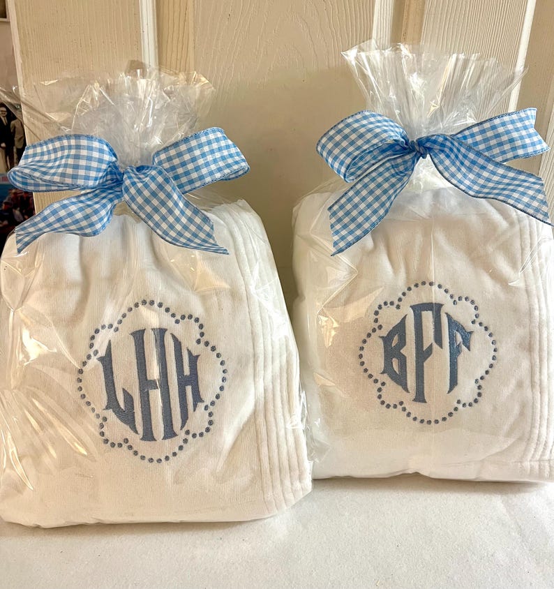 May include: Two white monogrammed gifts wrapped in clear cellophane and tied with blue and white gingham bows. Each gift features a different three-letter monogram in blue within a dotted scalloped border.