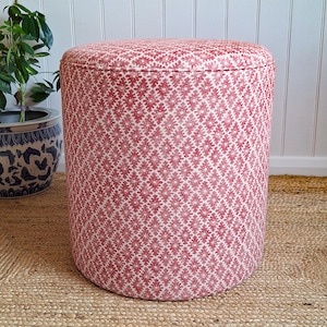 May include: A cylindrical ottoman upholstered in a red and white fabric featuring a repeating geometric floral pattern. The piece has a flat, circular top and straight sides, providing a stable structure for use as a seat or footrest.