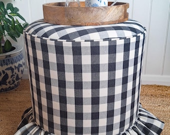 Könnte beinhalten: Ein zylindrischer Hocker, bezogen mit einem schwarz-weiß karierten Stoff im Büffel-Karo-Muster. Der untere Rand ist mit einer gerafften Stoffrüsche versehen. Auf dem Hocker steht ein rundes Holztablett mit einer kleinen Glasvase und Tasse.