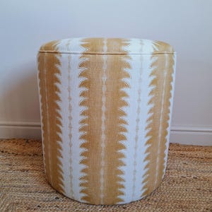 May include: A round ottoman with a white and ochre pattern. The fabric design features vertical rows of repeating, stylized leaf shapes. The ottoman sits on a woven, natural-fibre rug, and is against a plain white wall.