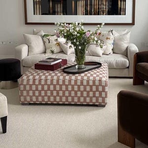 May include: A living room with a cream sofa, patterned cushions, and a red and cream checkered ottoman. A glass vase displays flowers. Other furnishings include a black footstool and brown armchairs. The room has a neutral colour scheme.