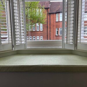 K&ouml;nnte beinhalten: Ein wei&szlig;er Fensterplatz mit einem hellgr&uuml;nen Kissen. Das Fenster hat wei&szlig;e Fensterl&auml;den und einen Blick auf ein Backsteingeb&auml;ude mit gr&uuml;nen B&auml;umen.