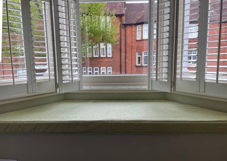 K&ouml;nnte beinhalten: Ein wei&szlig;er Fensterplatz mit einem hellgr&uuml;nen Kissen. Das Fenster hat wei&szlig;e Fensterl&auml;den und einen Blick auf ein Backsteingeb&auml;ude mit gr&uuml;nen B&auml;umen.