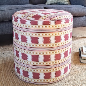 May include: A cylindrical ottoman upholstered in a woven fabric featuring a repeating pattern of horizontal bands in deep red, cream, and mustard yellow. The design includes alternating stripes and textured geometric blocks that wrap around the sides.