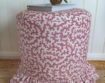 Könnte beinhalten: Ein zylindrischer Hocker, bezogen mit einem cremefarbenen Stoff mit einem sich wiederholenden dunkelroten, korallenartigen Muster. Die Basis des Hockers ist mit einer gerafften Stoffrüsche versehen, die nach außen verläuft und einen Rand bildet.